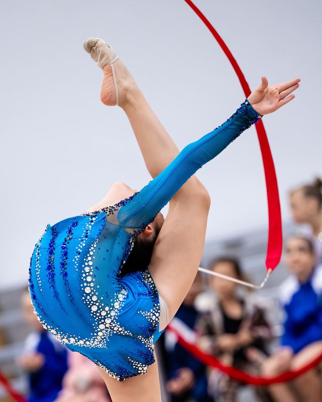 A gymnast mid-ribbon routine in a blue leotard on the competition floor
