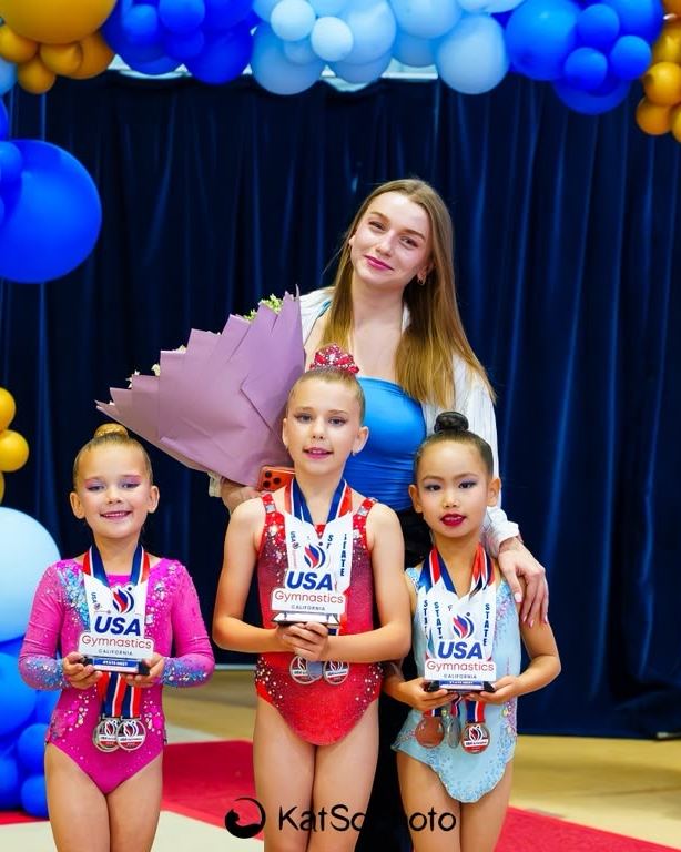 Three gymnasts holding their medals under a balloon arch with their coach