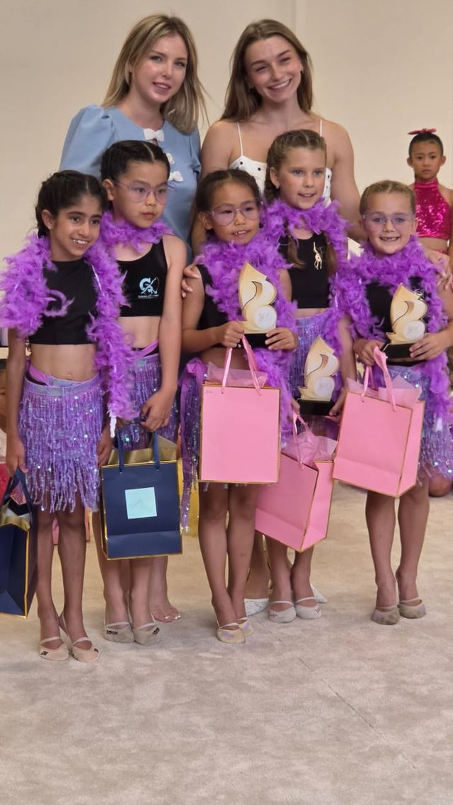 Five gymnasts in purple feather boas holding their show trophies